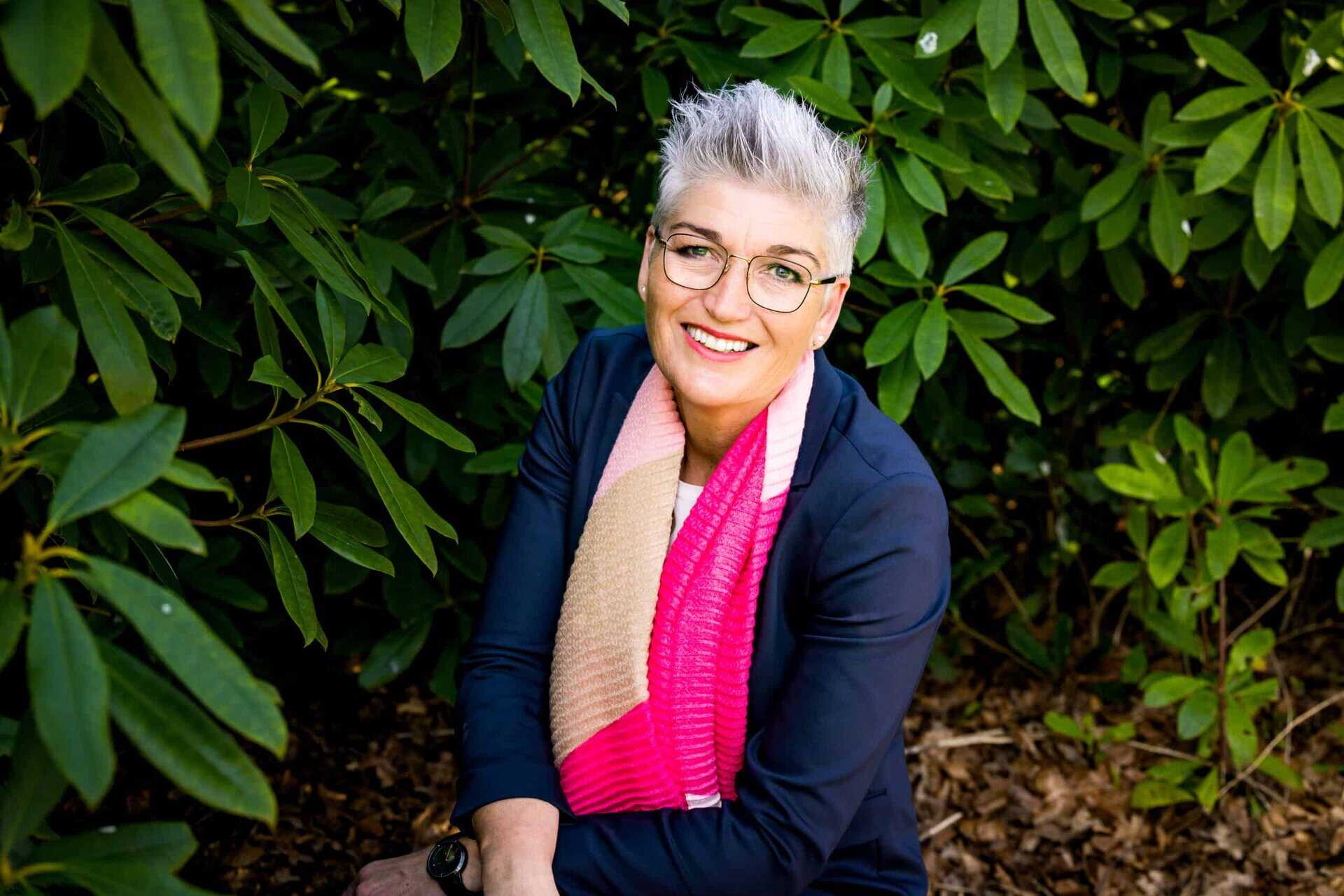 Natascha van Zeijl die met een bril en een roze sjaal in een struik zit, kijkt uit over Natascha.
